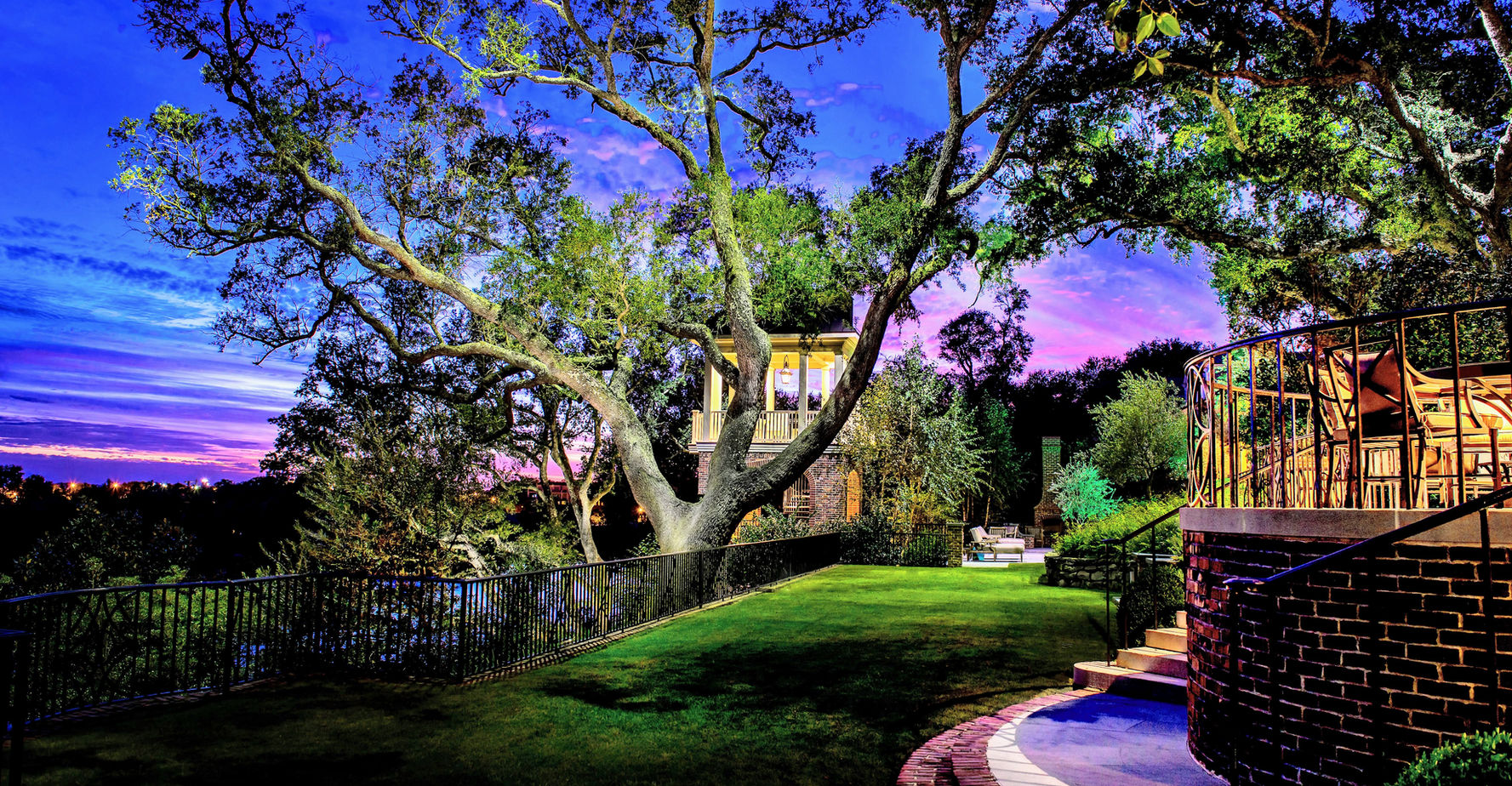 A large tree and modern house with outdoor lights stand on a well-manicured lawn at dusk, with a vibrant purple and blue sky in the background.