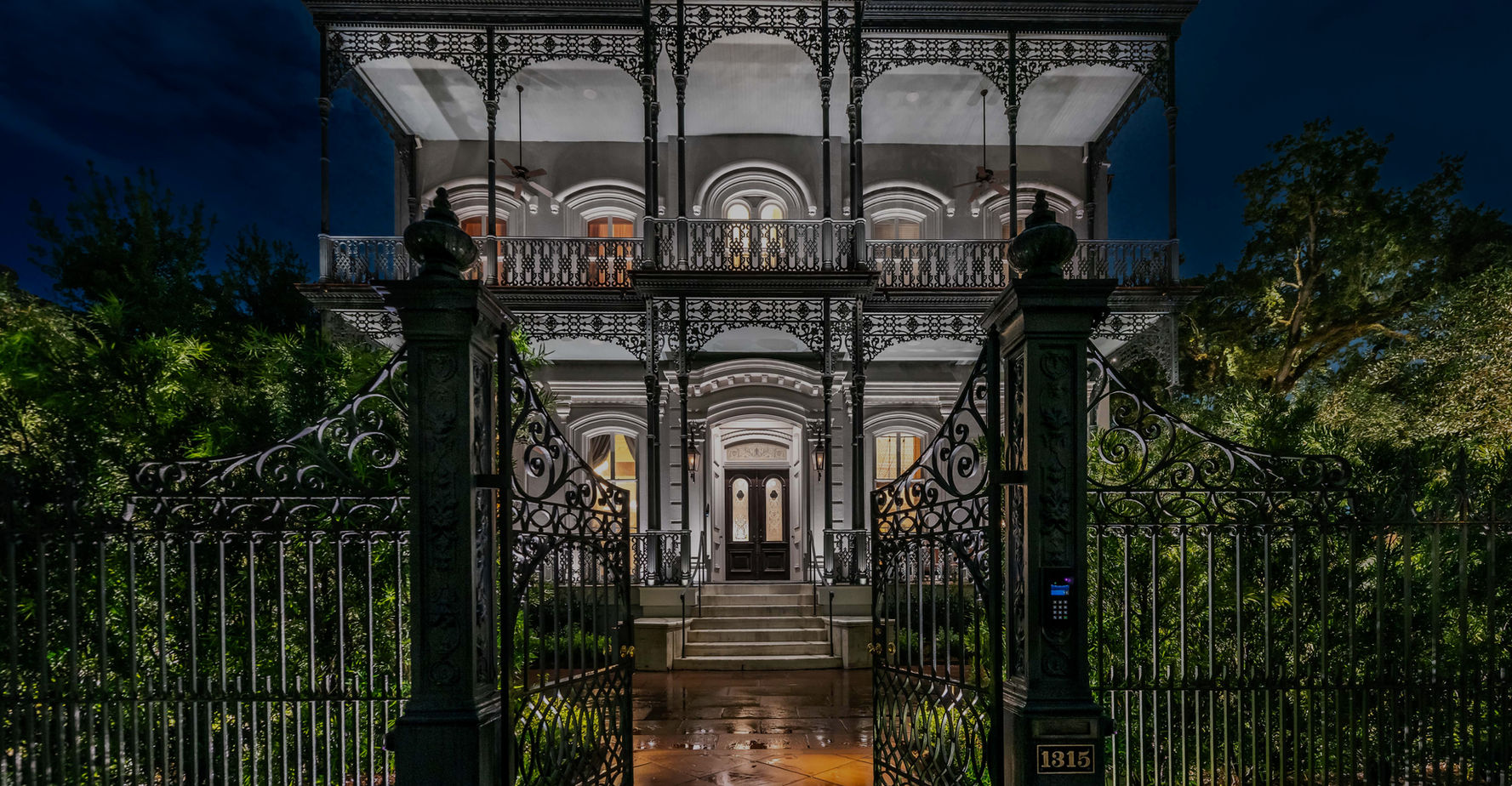 A large, two-story house with ornate ironwork, arched windows, and a wrought-iron gate, illuminated at night.