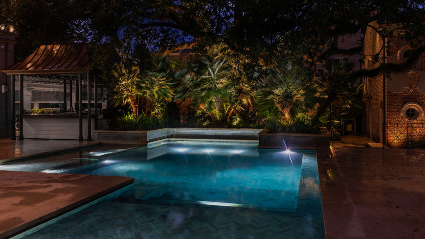 A swimming pool at night is illuminated by underwater lights, with surrounding palm trees and foliage highlighted by landscape lighting.