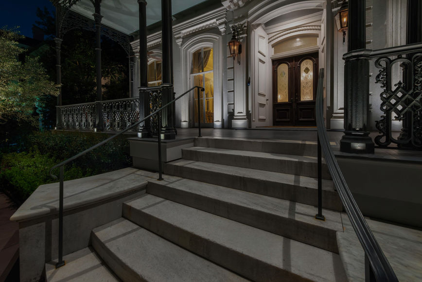 Wide stone steps lead up to the front entrance of a large, well-lit house with columns, ornate railings, and double doors at night.