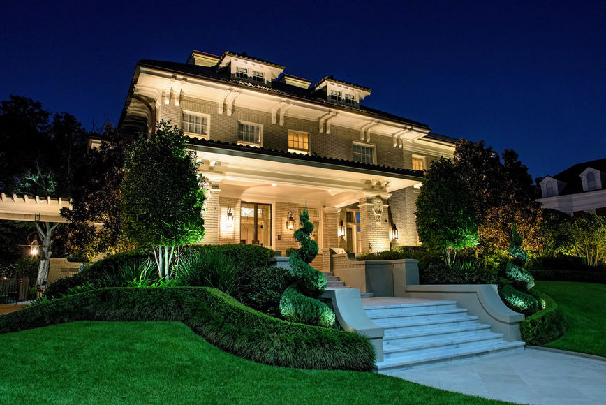 Large, well-lit two-story house with manicured landscaping, multiple windows, and a wide staircase leading to the front entrance at night.