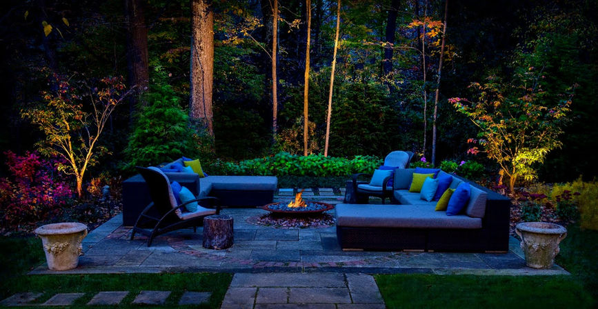 Outdoor patio with modern furniture, cushions, and a fire pit, surrounded by trees and plants, illuminated by colorful landscape lighting at night.