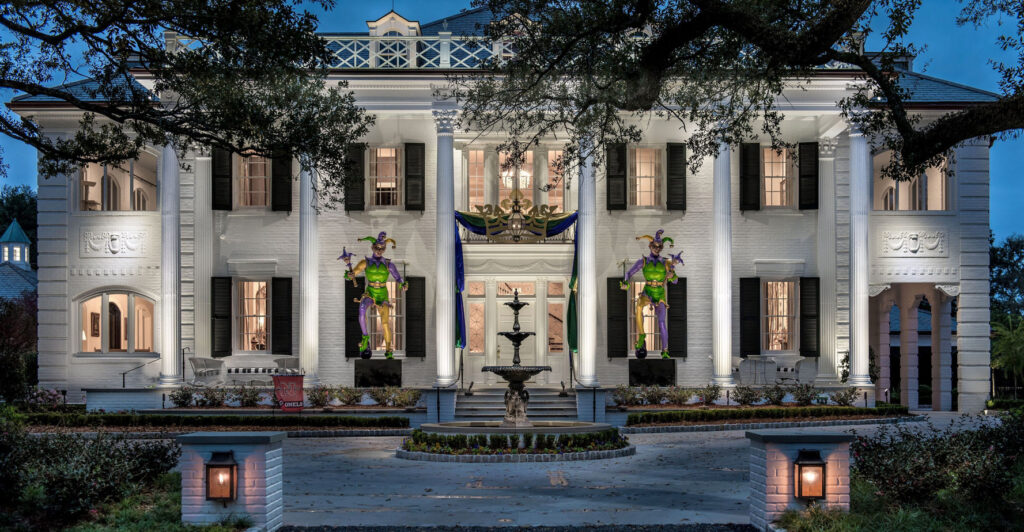 Large white mansion with columns, black shutters, and festive Mardi Gras jester decorations on the front porch, lit up in the evening. Fountain and landscaped garden in the foreground.