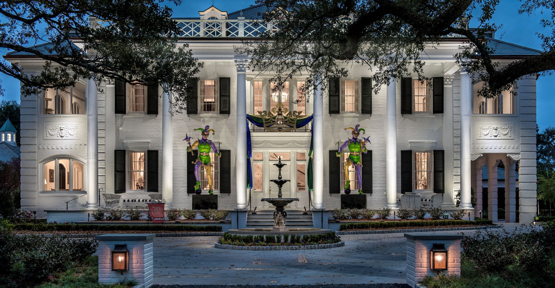 Large white mansion with columns, black shutters, and festive Mardi Gras jester decorations on the front porch, lit up in the evening. Fountain and landscaped garden in the foreground.