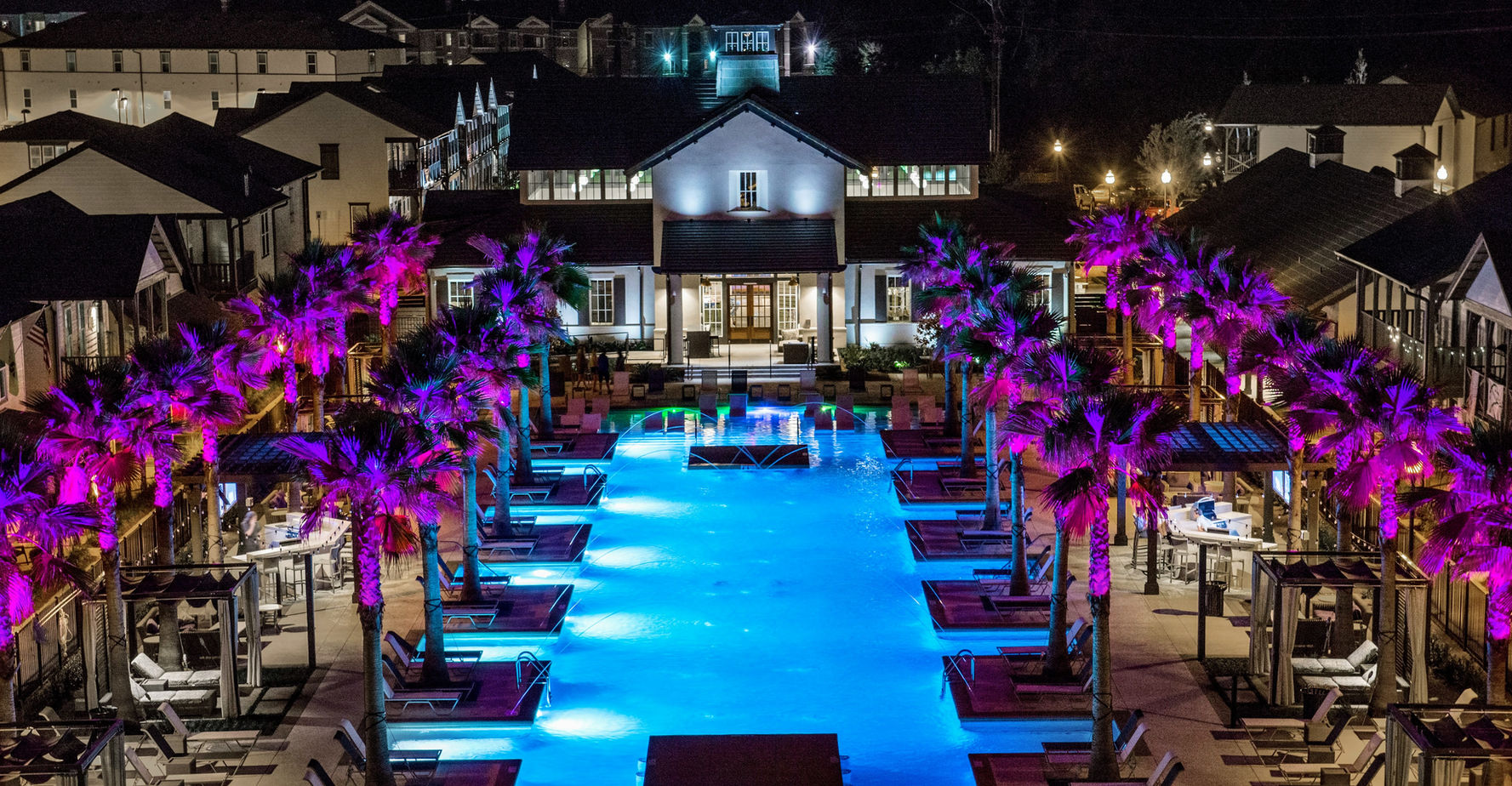 Night view of a large illuminated swimming pool surrounded by lounge chairs and palm trees with purple lighting in a residential complex.