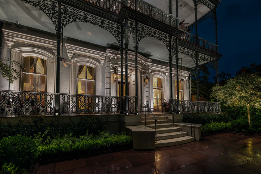 A historic building with ornate iron railings, large windows, and exterior lighting, seen at night with greenery along the walkway.