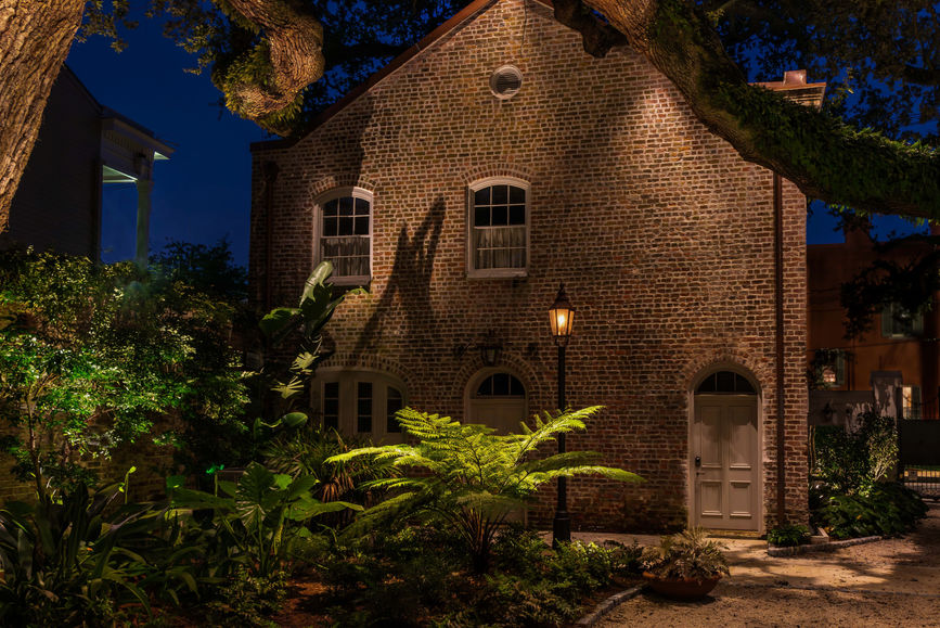 A brick house with arched windows and doors is illuminated by a streetlamp at night, surrounded by lush greenery and shaded by large tree branches.