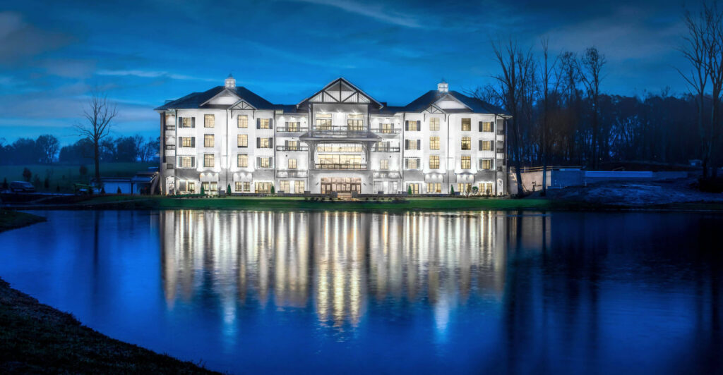 A large, well-lit building with multiple windows is reflected in a calm pond at dusk, surrounded by trees and a grassy area.