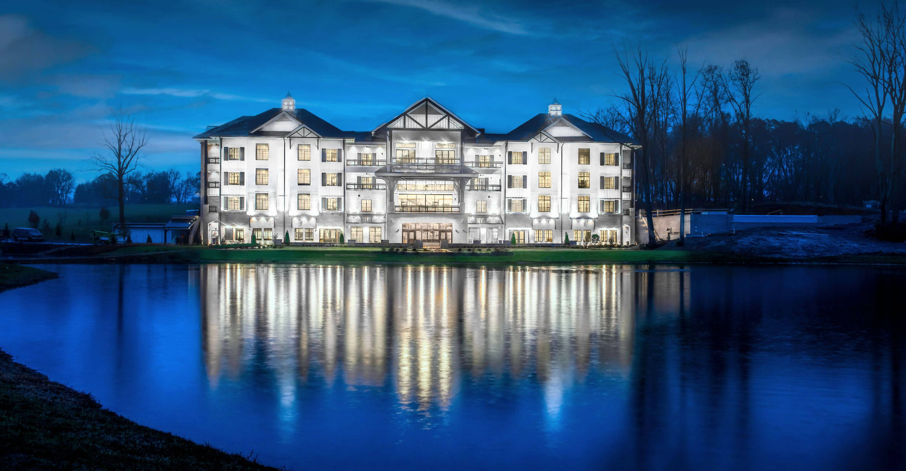 A large, well-lit building with multiple windows is reflected in a calm pond at dusk, surrounded by trees and a grassy area.