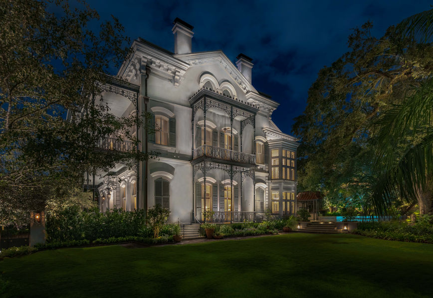 A large, ornate, two-story white mansion with balconies, decorative railings, and columns, illuminated at night, surrounded by trees and a manicured lawn.