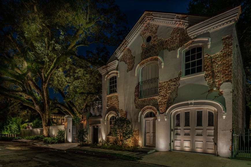 A two-story house with a mix of exposed brick and stucco exterior, arched garage doors, and nighttime landscape lighting highlighting trees and facade.