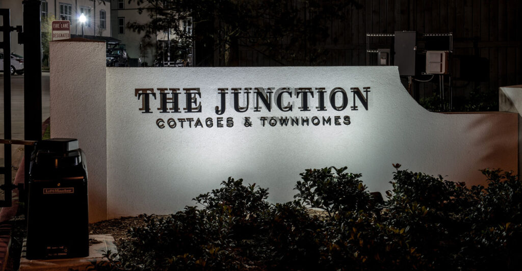 A white sign reads "The Junction Cottages & Townhomes," illuminated at night with plants in front and nearby buildings visible in the background.
