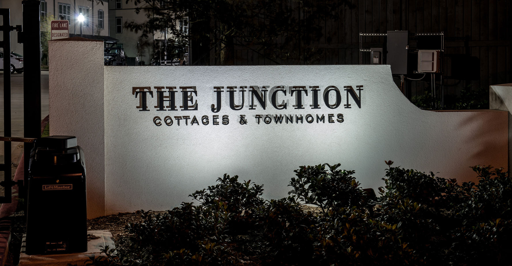 A white sign reads "The Junction Cottages & Townhomes," illuminated at night with plants in front and nearby buildings visible in the background.