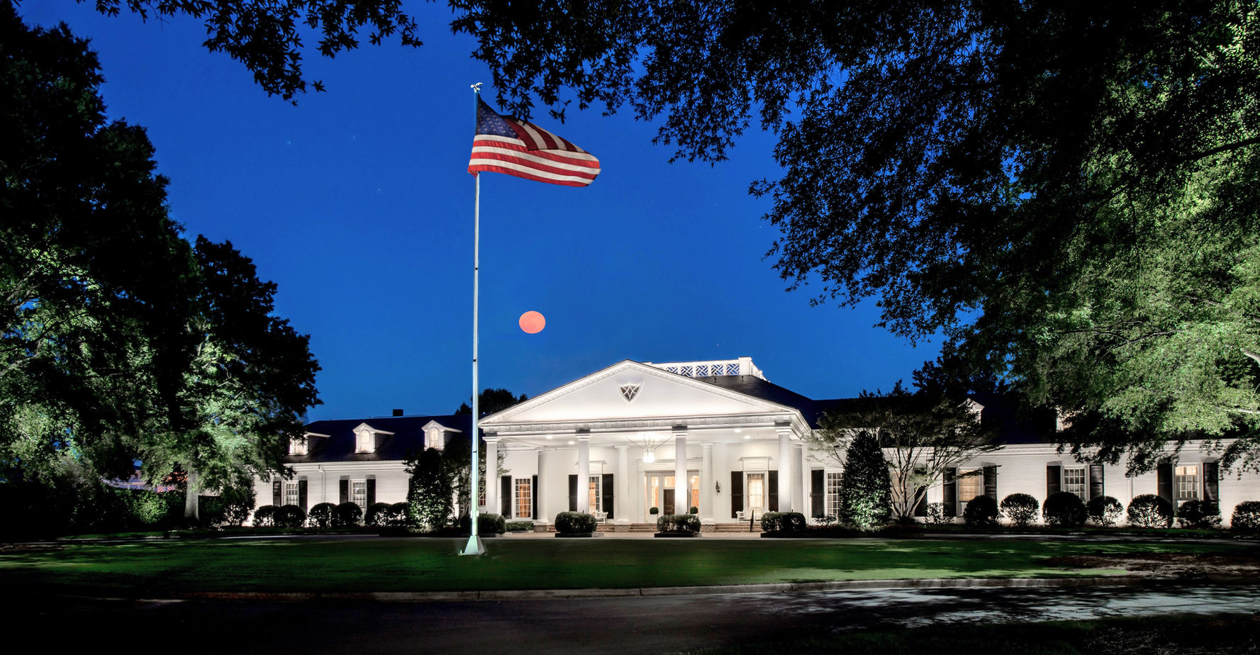 A large white building with columns is illuminated at night, with an American flag on a tall pole in the front lawn and a red moon in the sky.