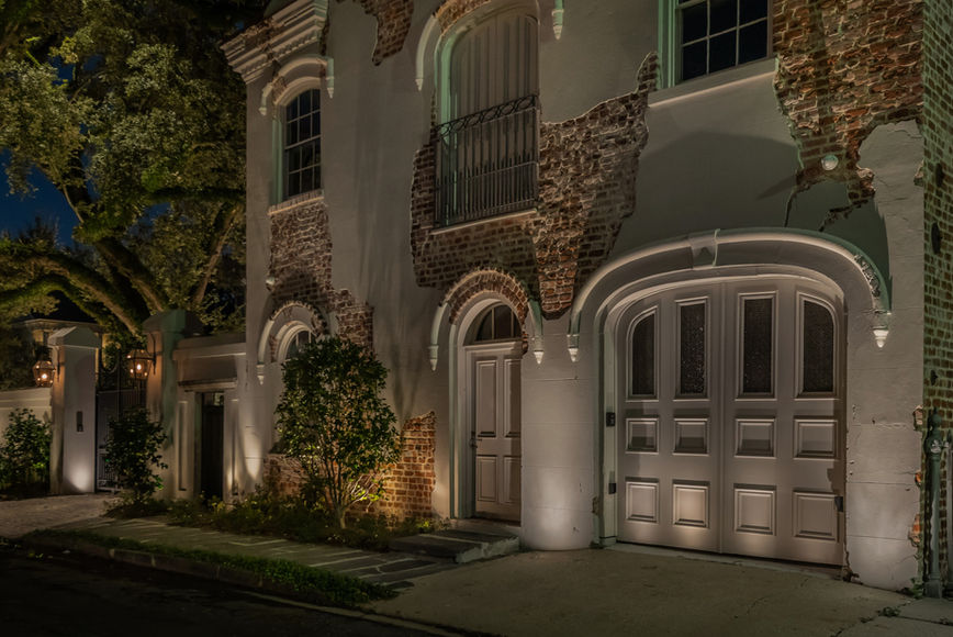 A historic brick and stucco building at night with arched garage doors, a balcony, and spotlights illuminating the façade.