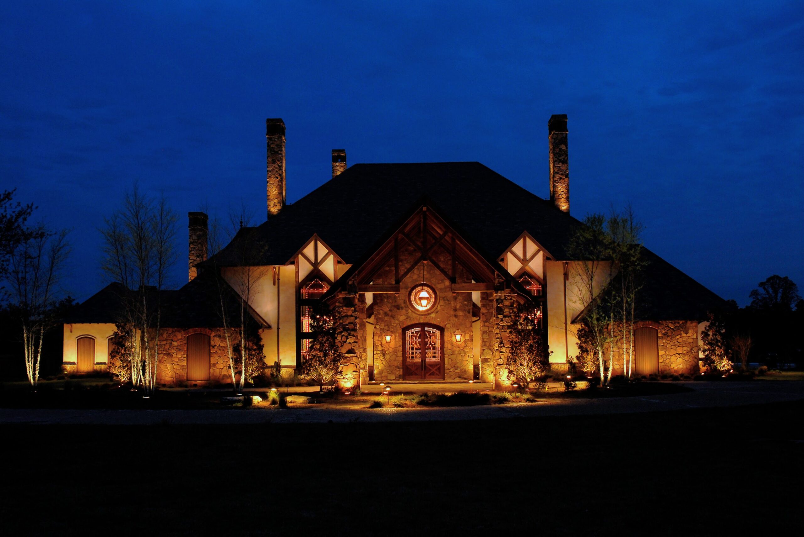 Large, well-lit stone house with a steep roof, multiple chimneys, and decorative wooden elements, photographed at dusk against a dark blue sky.