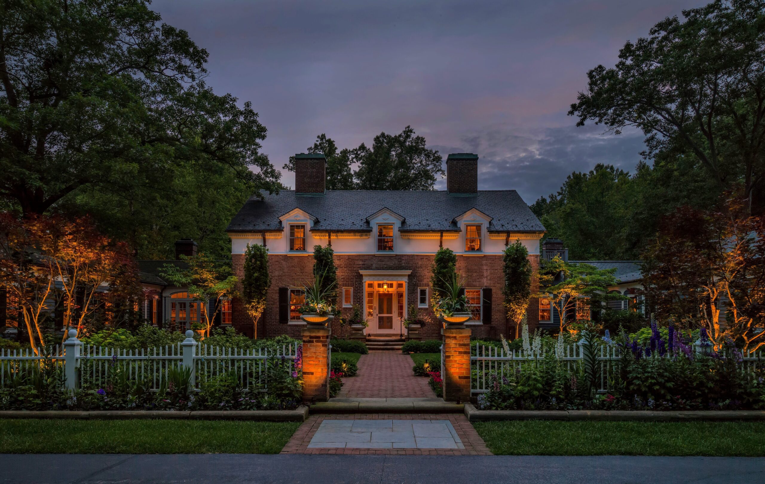 A large brick house with lit windows is surrounded by trees, landscaped gardens, and a white picket fence at dusk.