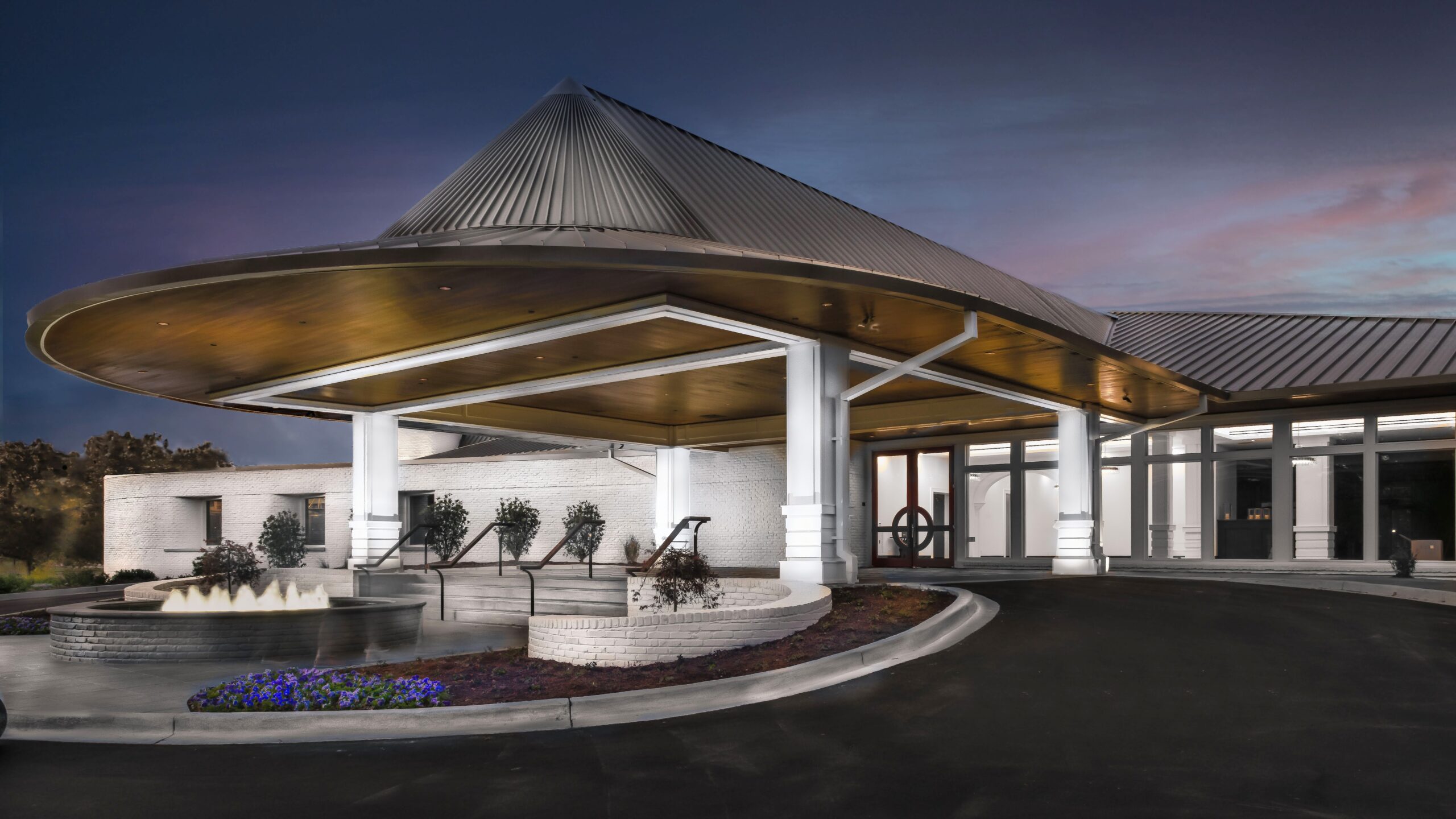 Modern building with a metal roof and covered entrance, featuring white columns, a small circular fountain, and landscaped flower beds in the foreground at dusk.