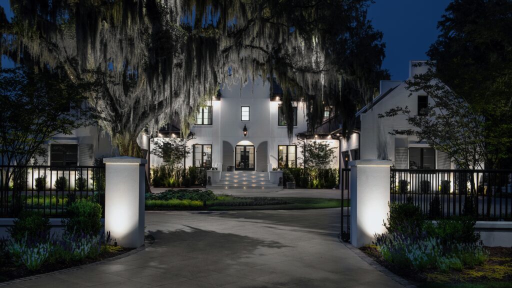 A large white house with multiple windows is illuminated at night, viewed through an open gate with trees and hanging moss in the foreground.