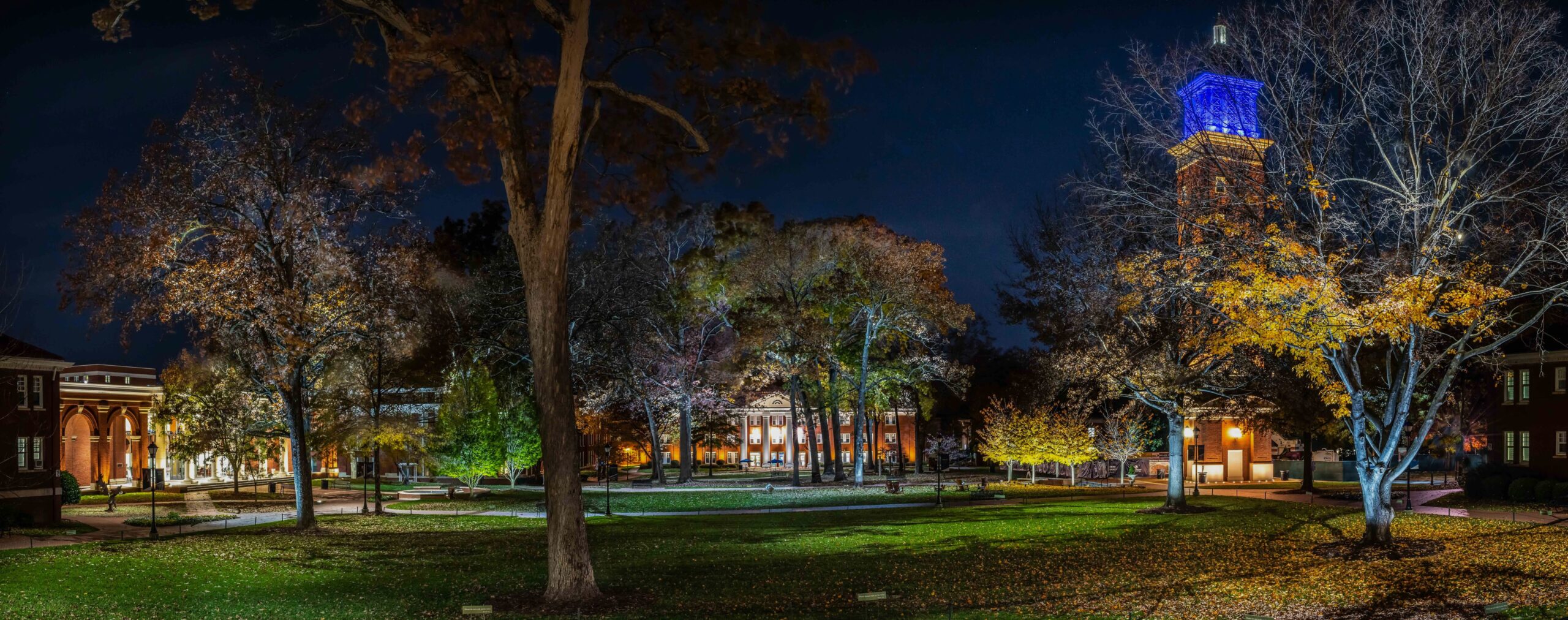 A nighttime view of a college campus quad with illuminated buildings and a tall clock tower with blue lighting, surrounded by trees and grass.