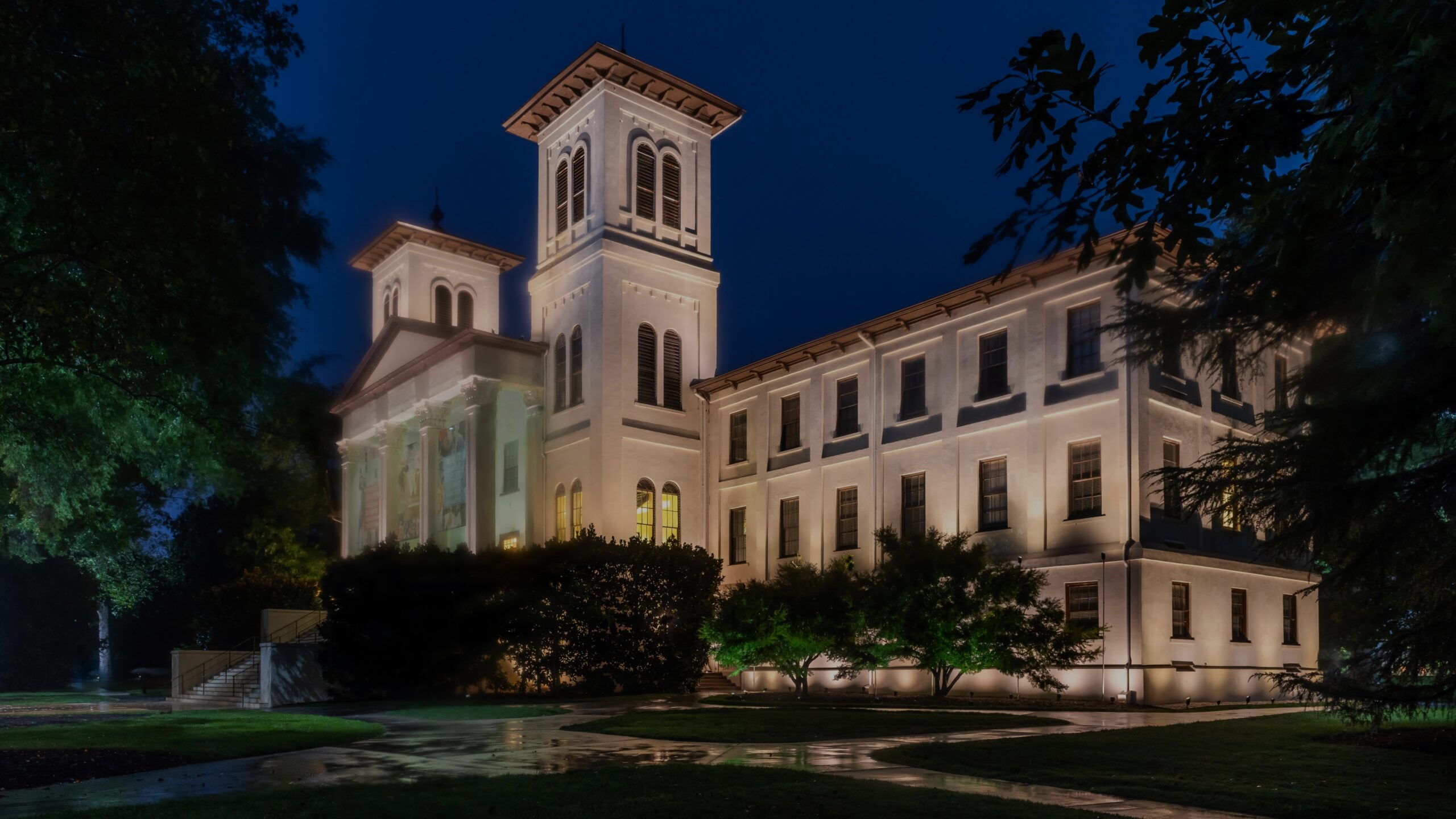 Historic building with tall towers and illuminated exterior at night, surrounded by trees, with wet pavement in the foreground.